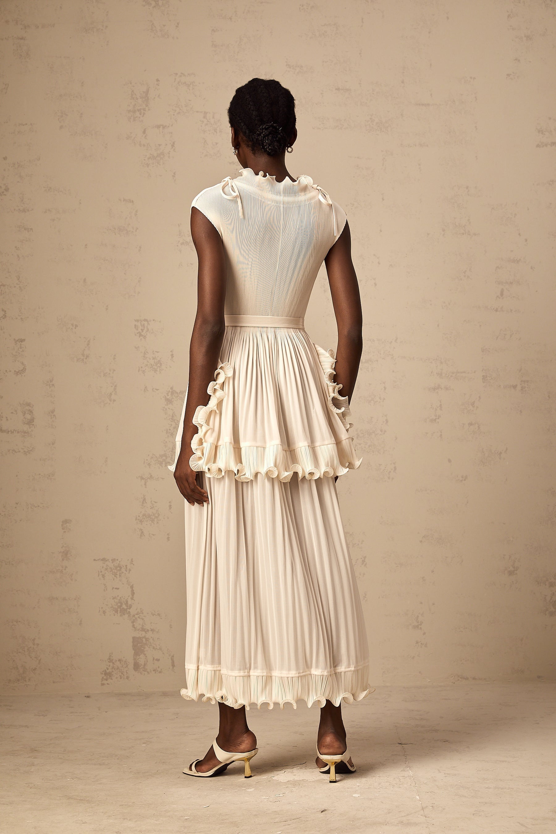 A woman wearing a long white sleeveless maxi dress with ruffle details and pleats standing against a plain background