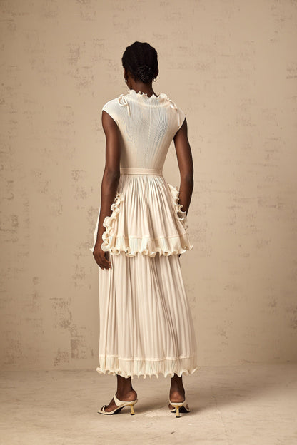 A woman wearing a long white sleeveless maxi dress with ruffle details and pleats standing against a plain background