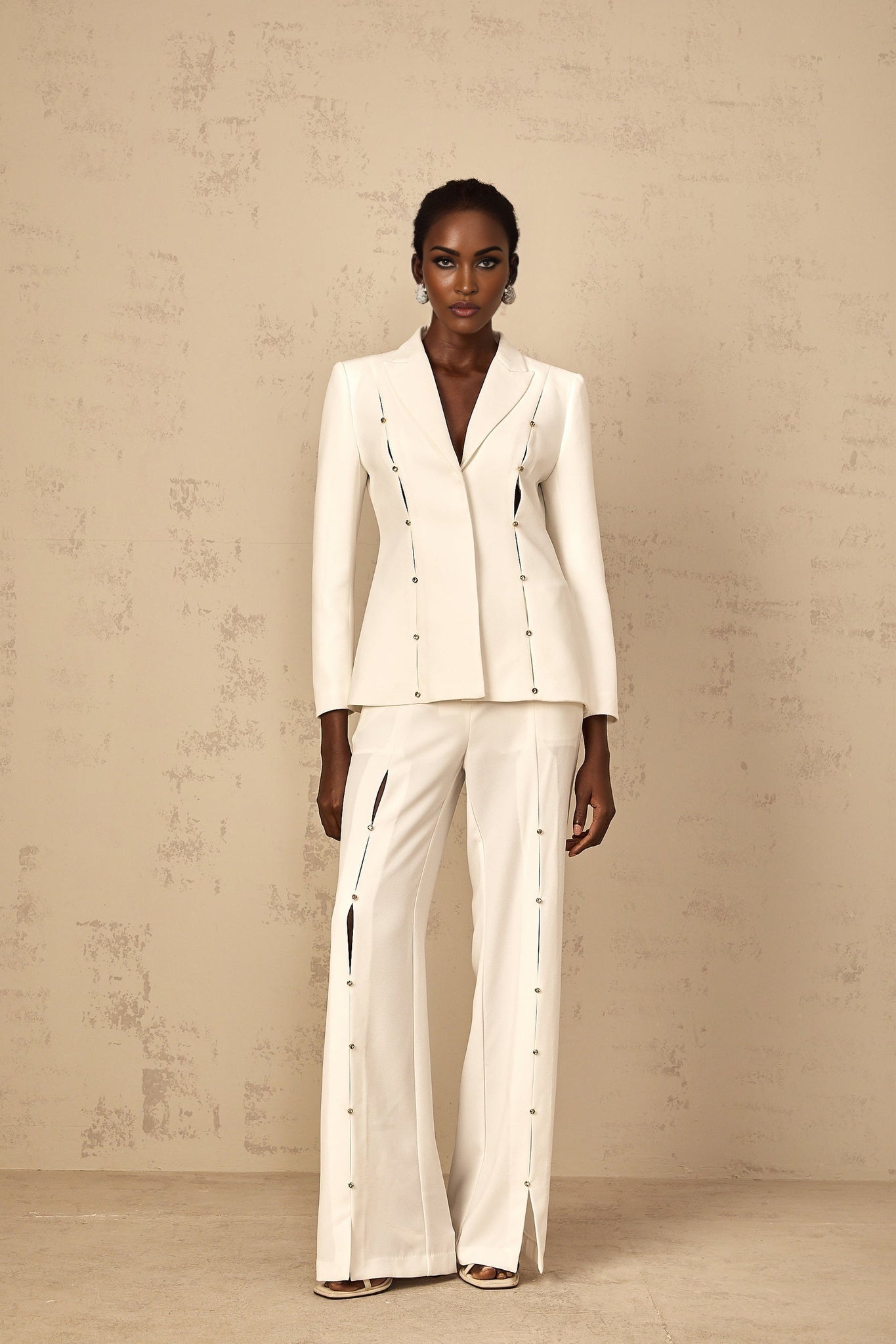 A woman wearing a matching blazer and trouser pant set in a light color The blazer features embellishments and the outfit has a tailored fit named Frida