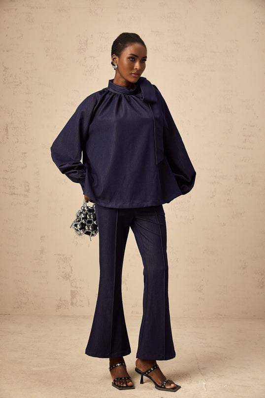 A woman wearing a matching denim blue outfit consisting of a flared blouse and trousers The blouse has long sleeves and the trousers have a wide leg silhouette The set is called Mathilde and has a casual yet stylish appearance