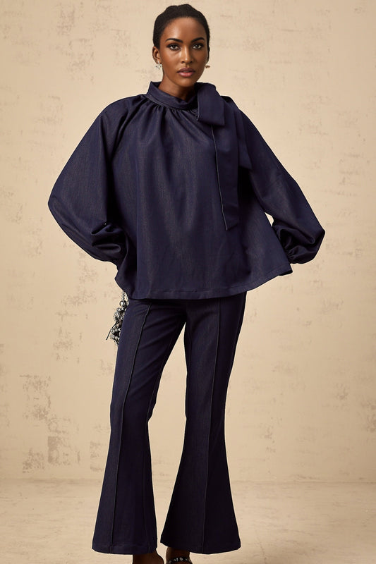 A woman wearing a matching denim blue outfit featuring a flared blouse and trousers standing against a plain background