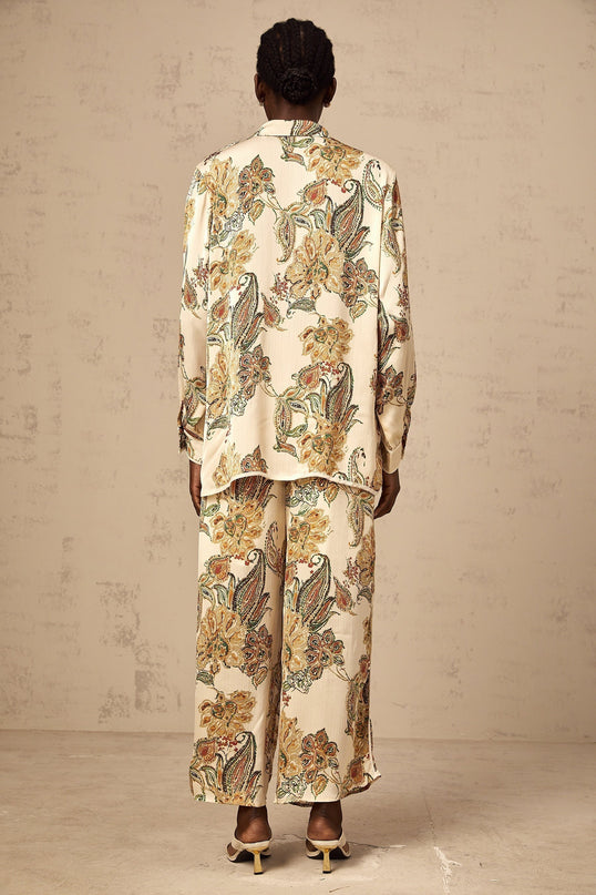 A woman wearing a matching floral print shirt and trousers set in neutral colors featuring an allover floral pattern The outfit has a relaxed fit and elegant style