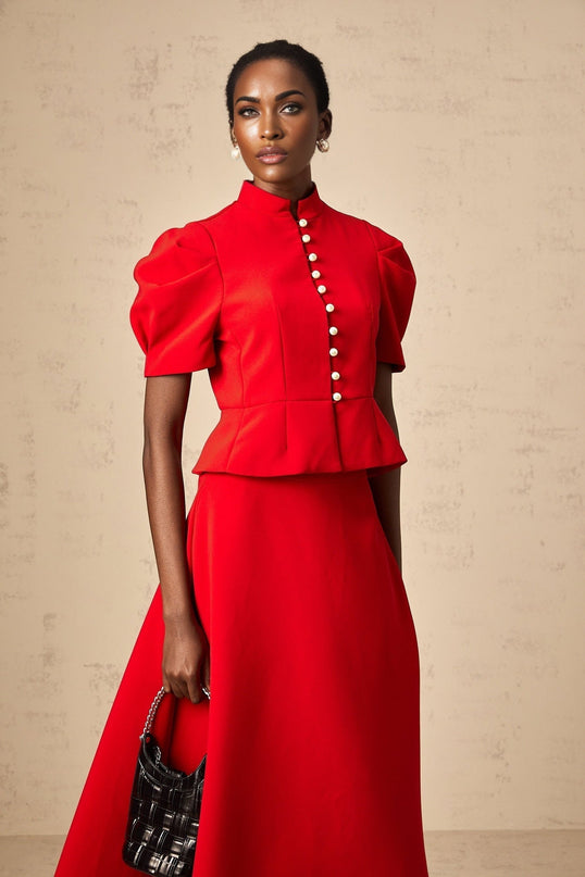 A woman wearing a matching red outfit consisting of a pleated blouse and skirt the blouse has a high collar and the skirt falls to mid calf length