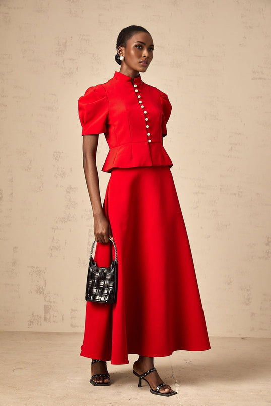 A woman wearing a matching red outfit consisting of a pleated blouse and skirt The blouse has a high neck and short sleeves while the skirt falls to about knee length Both pieces feature pleated fabric in a vibrant red color