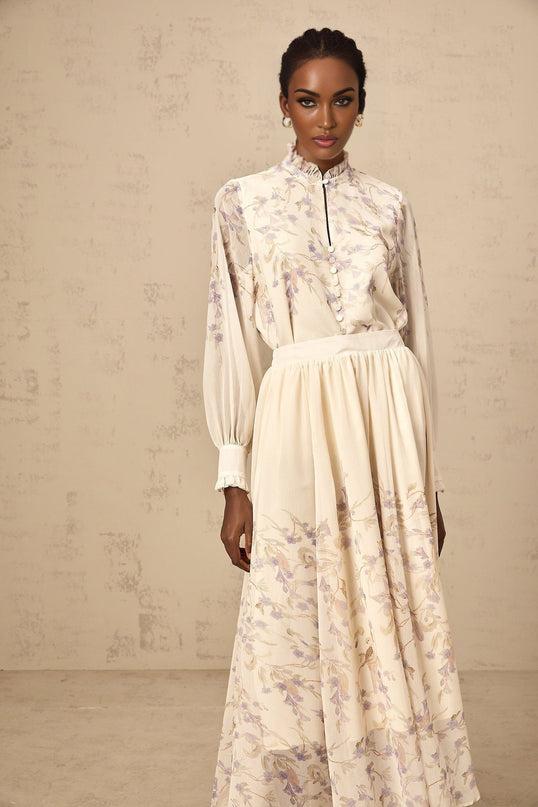 A woman wearing a matching set consisting of a frilled wisteria print shirt and skirt in a light purple color The shirt has ruffled details and the skirt falls to mid length creating an elegant and coordinated outfit