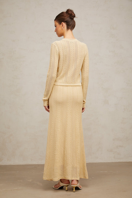A woman wearing a matching set consisting of a pointelleknit cardigan and skirt in a light color The cardigan has long sleeves and an open front while the skirt falls to mid length The set is named Tiphaine and features an intricate knit pattern