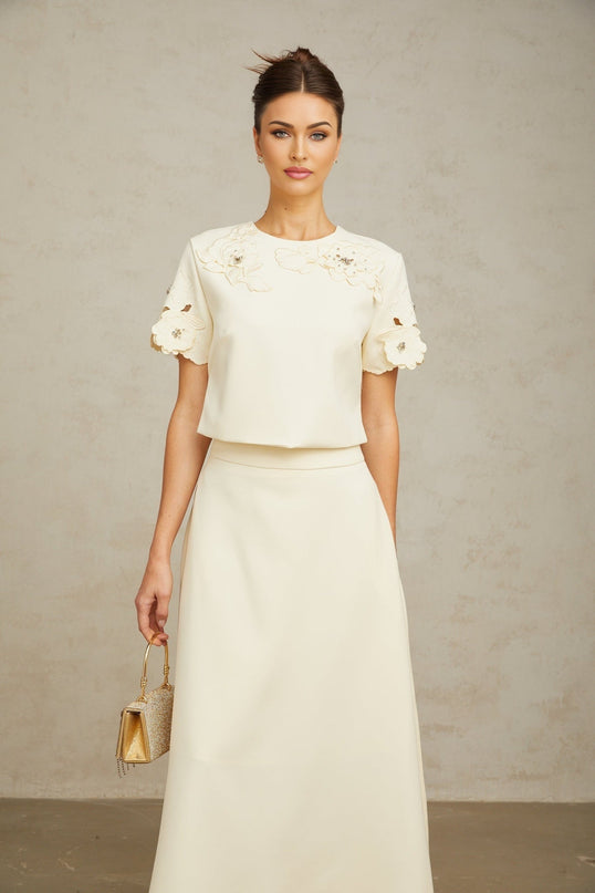 A woman wearing a matching set consisting of a sleeveless top and skirt in neutral tones featuring three dimensional petal details covering the fabric