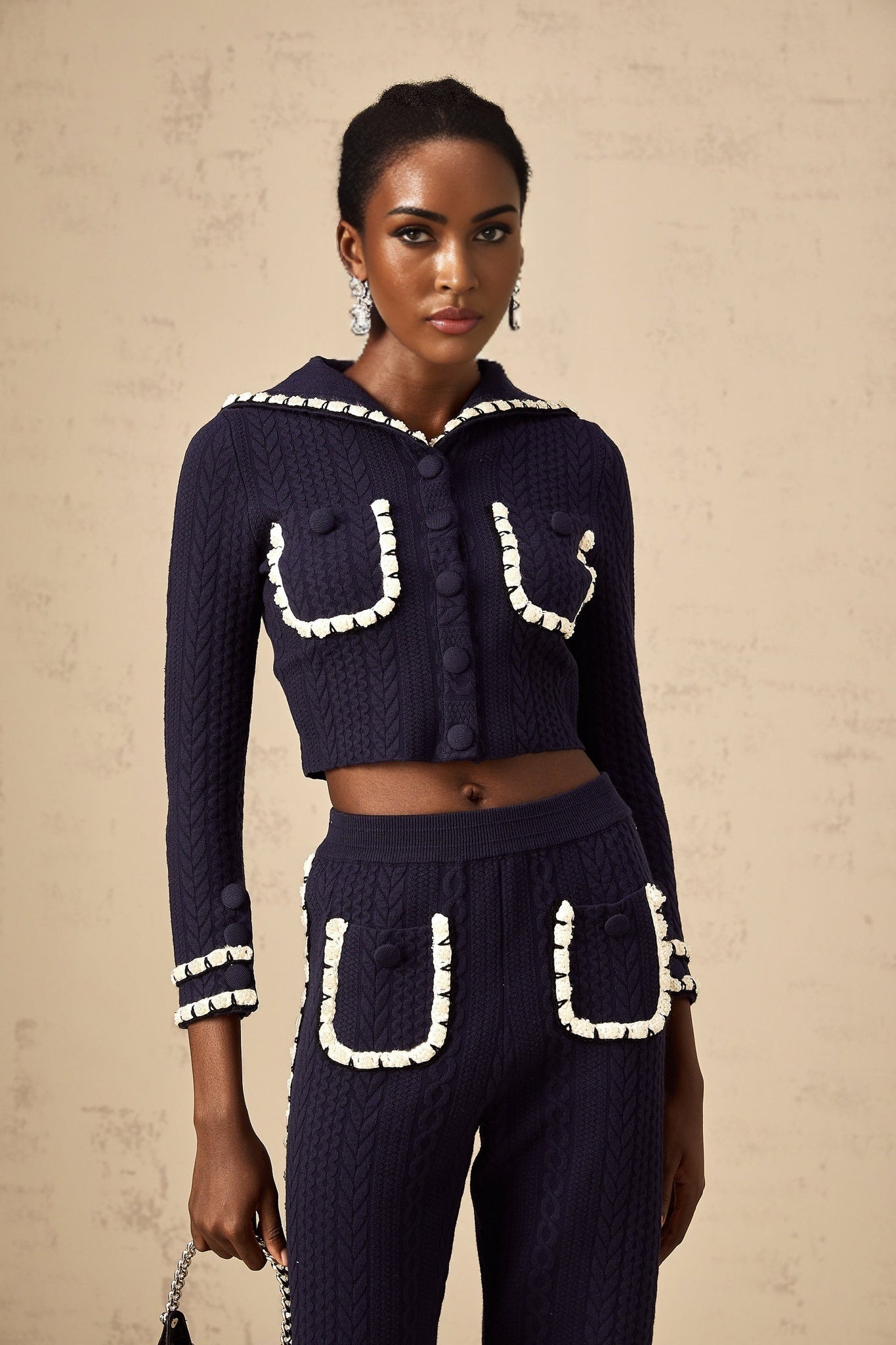 A woman wearing a navy blue crochet knit cardigan and matching trousers standing against a plain background