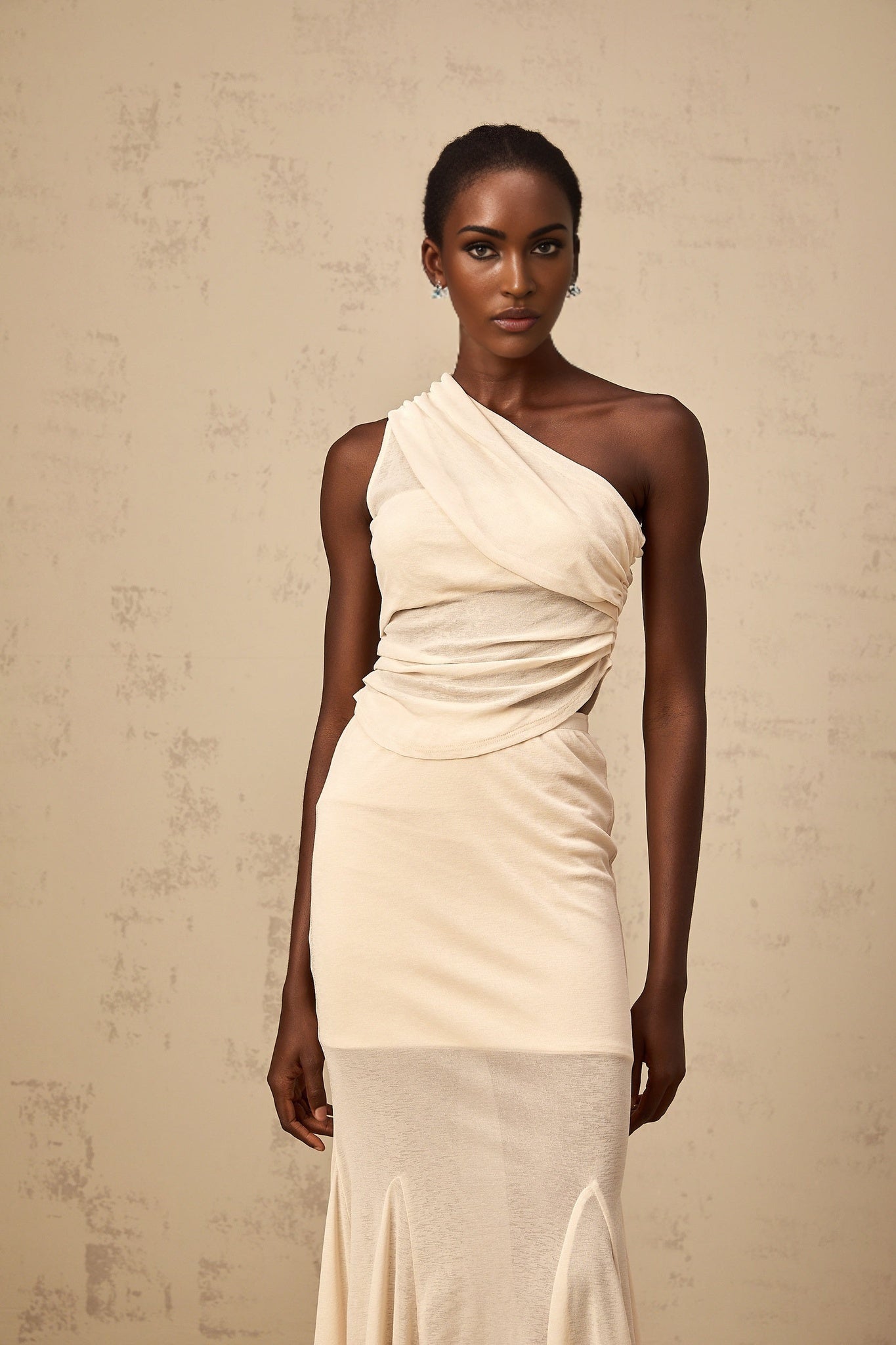 A woman wearing a neutralcolored matching set consisting of a ruched stretch top and skirt The top has long sleeves and the skirt is formfitting Both pieces feature gathered fabric details