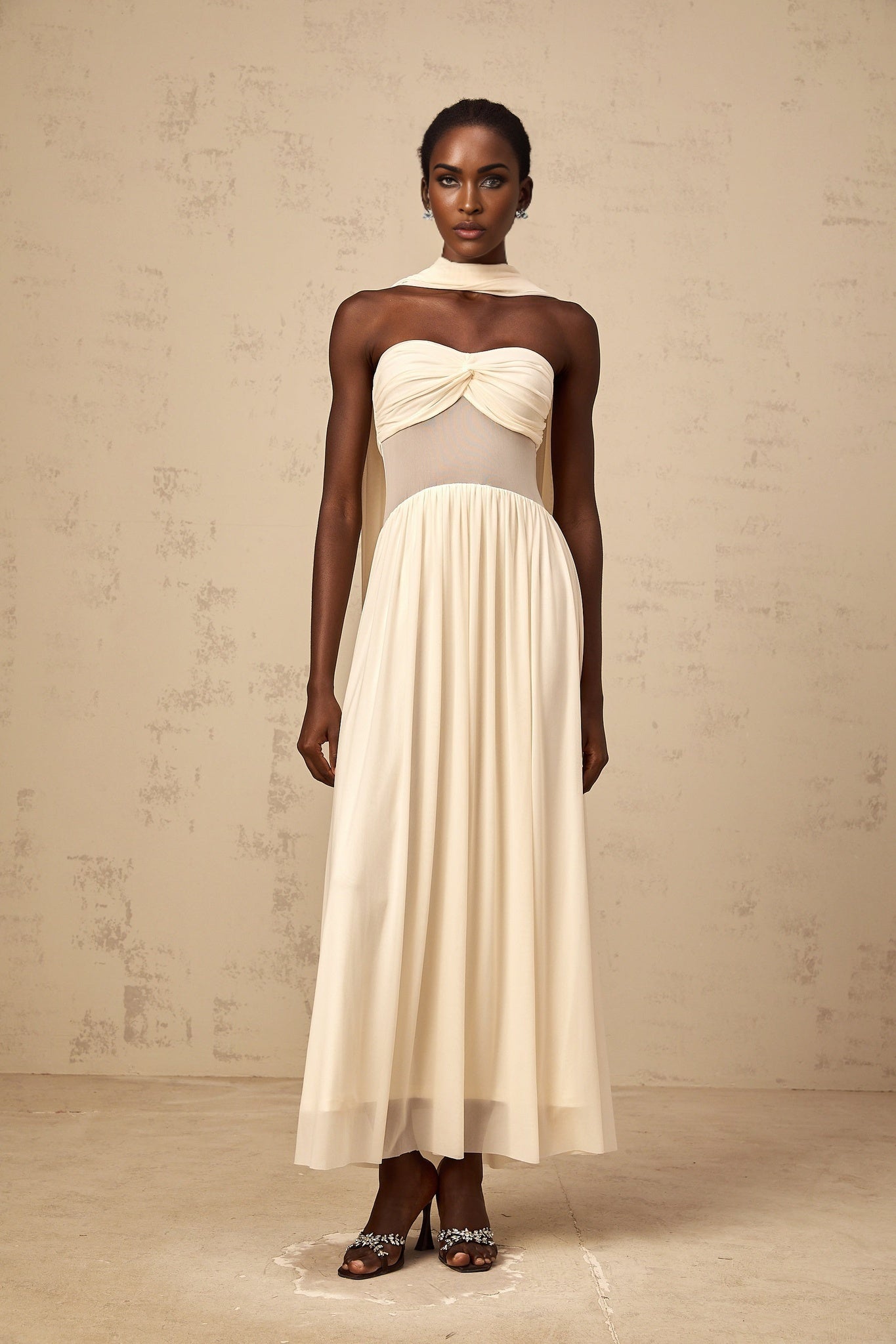 A woman wearing a neutralcolored midi dress with a scarfinspired mesh design featuring flowy fabric and intricate patterns