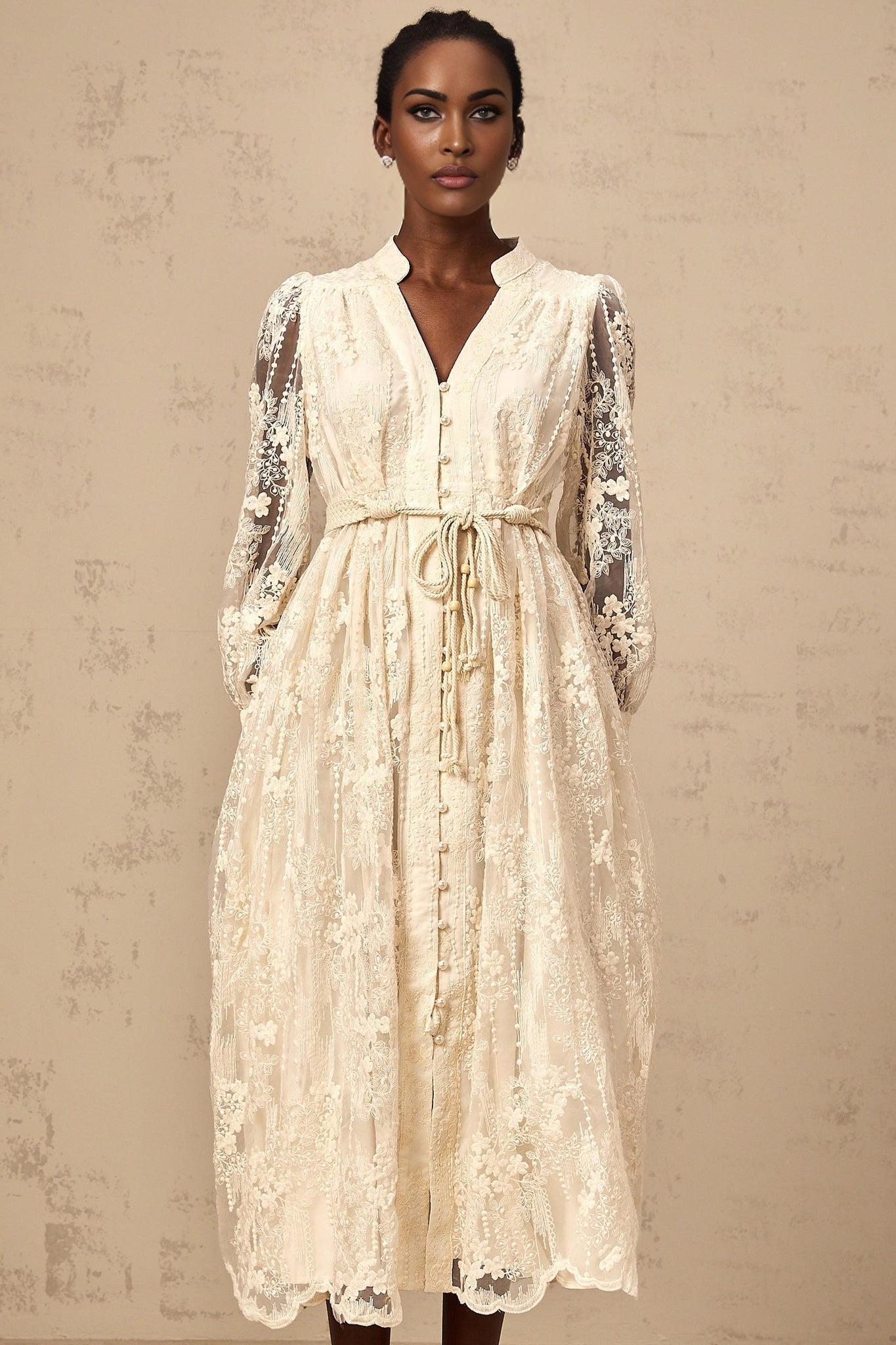 A woman wearing a neutralcolored midi dress with lace embroidery detailing standing in a relaxed pose against a plain background
