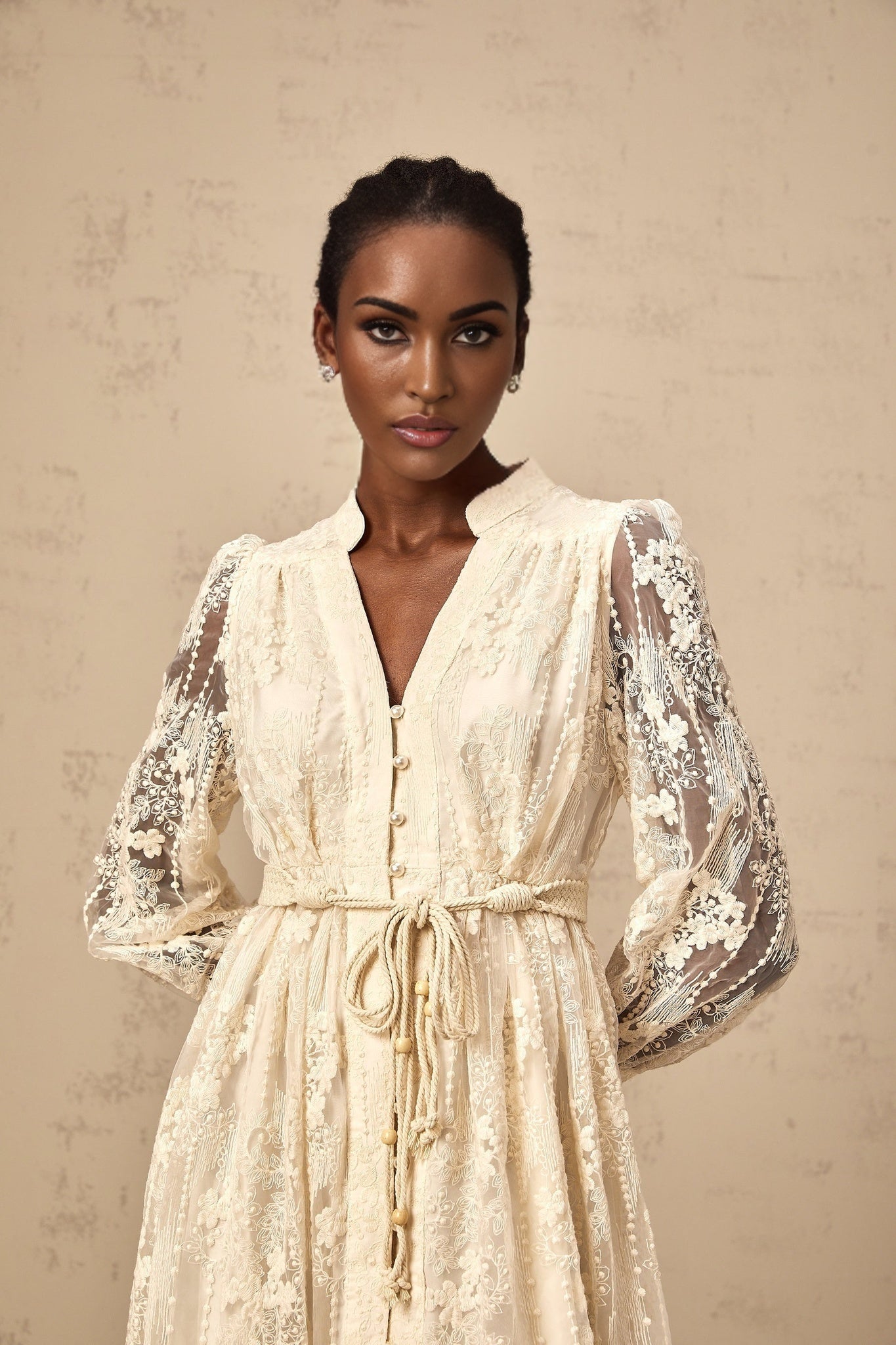 A woman wearing a neutralcolored midi dress with lace embroidery detailing standing in a relaxed pose against a plain background