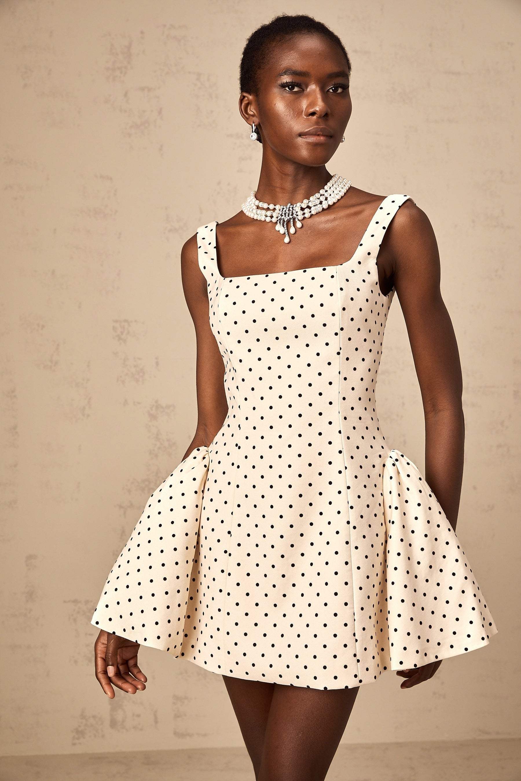 A woman wearing a neutralcolored mini dress with polka dots and puff sleeves standing in a casual pose