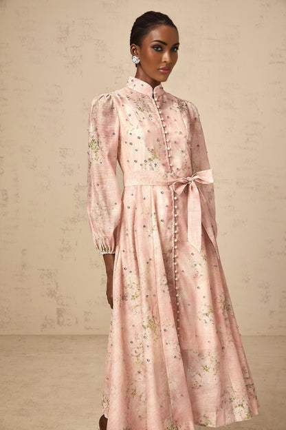A woman wearing a pink midi dress with floral print and embellishments standing against a plain background