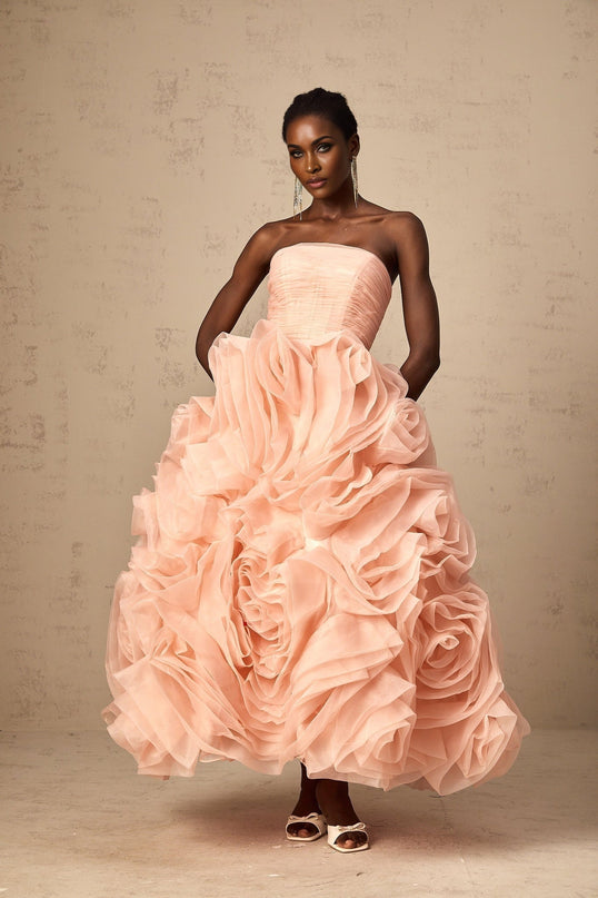 A woman wearing a pink offshoulder midi dress with 3D petal details on the tulle fabric standing in a neutral background