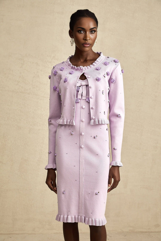 A woman wearing a purple knitted cardigan and matching dress adorned with faux flower details standing against a plain background