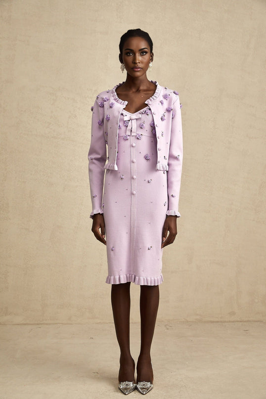 A woman wearing a purple knitted cardigan and matching dress with floral embellishments standing against a plain background