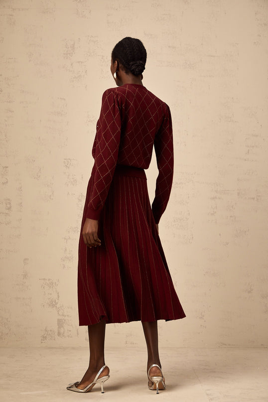 A woman wearing a red knit cardigan and matching skirt set with argyle pattern and embellishments The cardigan features long sleeves and buttons while the skirt is knee length Both pieces have an intarsia knit design in red with argyle detailing