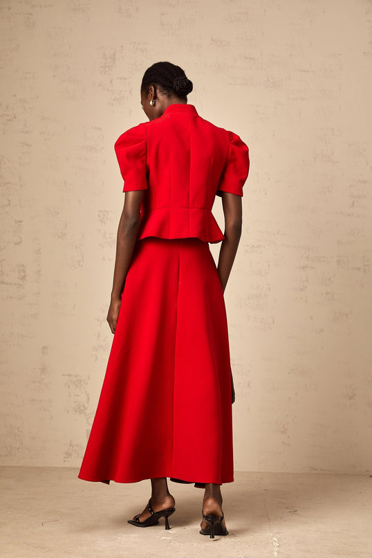 A woman wearing a red matching set consisting of a pleated blouse and skirt The blouse has long sleeves and the skirt falls to mid thigh length The outfit is styled with black heels