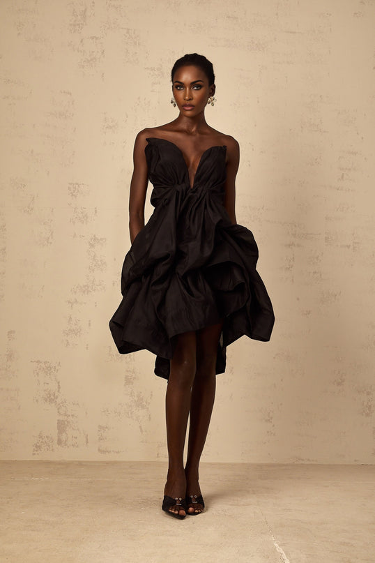 A woman wearing a short asymmetrical cocktail dress with pleated details The dress has one shoulder and falls above the knee The model is shown in a standing pose against a plain background