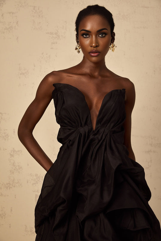 A woman wearing a short asymmetrical pleated cocktail dress named Elida The dress features a one shoulder design with a fitted bodice and a flared skirt that falls above the knee