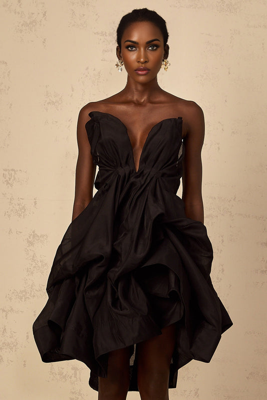 A woman wearing a short asymmetrical pleated cocktail dress with one shoulder strap The dress has a fitted bodice and flared skirt in a vibrant color