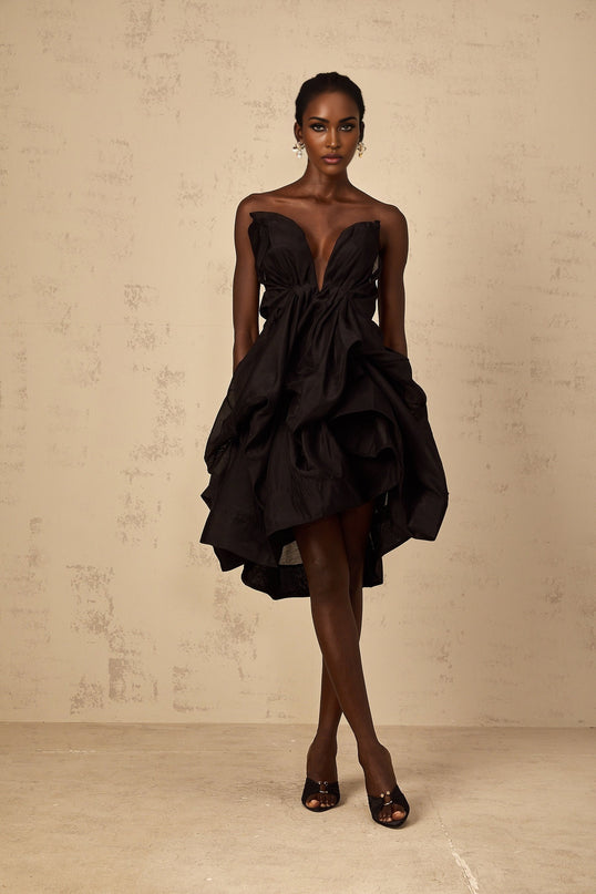 A woman wearing a short asymmetrical pleated cocktail dress with one shoulder strap The dress has a fitted bodice and flared skirt in a vibrant color