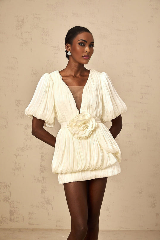 A woman wearing a short beige dress with pleats and floral appliques standing in a neutral setting