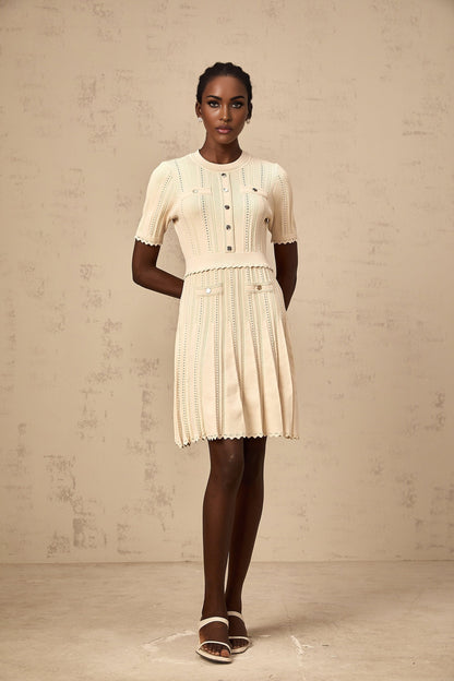 A woman wearing a short beige knitted dress with pleated skirt and delicate pointelle trim standing in a neutral background