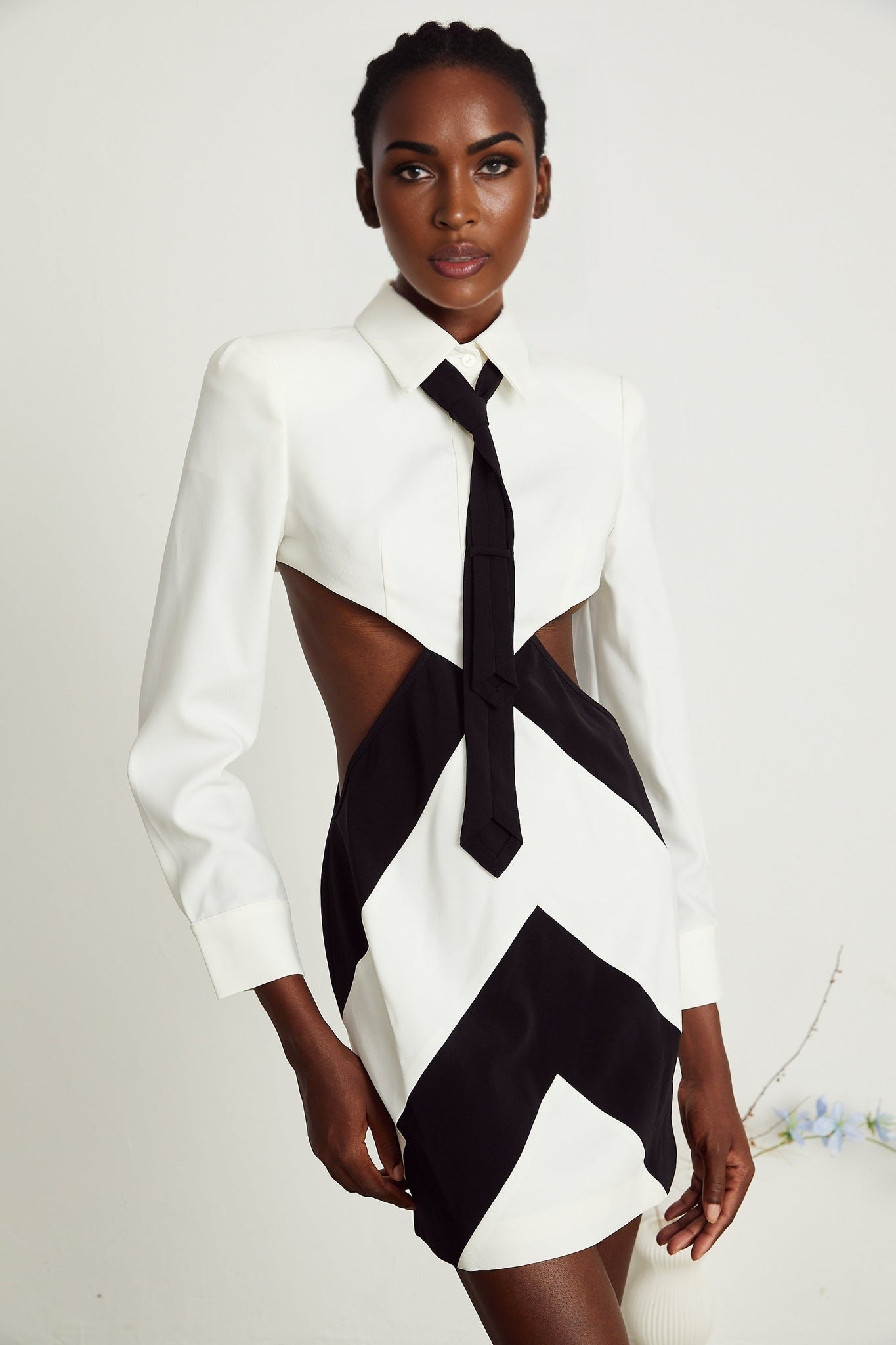 A woman wearing a short black dress with a halter neckline and tie detail The dress features a cutout design revealing some skin on the torso The model stands in a relaxed pose showing off the dresses fitted silhouette