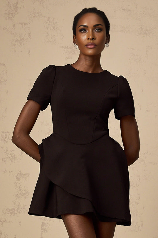 A woman wearing a short black dress with an asymmetric neckline and ruffled details standing against a plain background