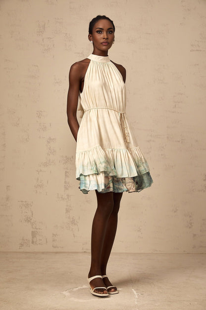 A woman wearing a short multicolored layered dress with a mixed pattern print standing against a plain background