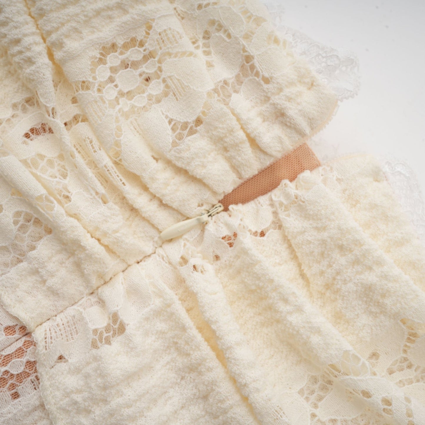A woman wearing a short neutral colored floral lace mini dress with flared cuffs standing against a plain background