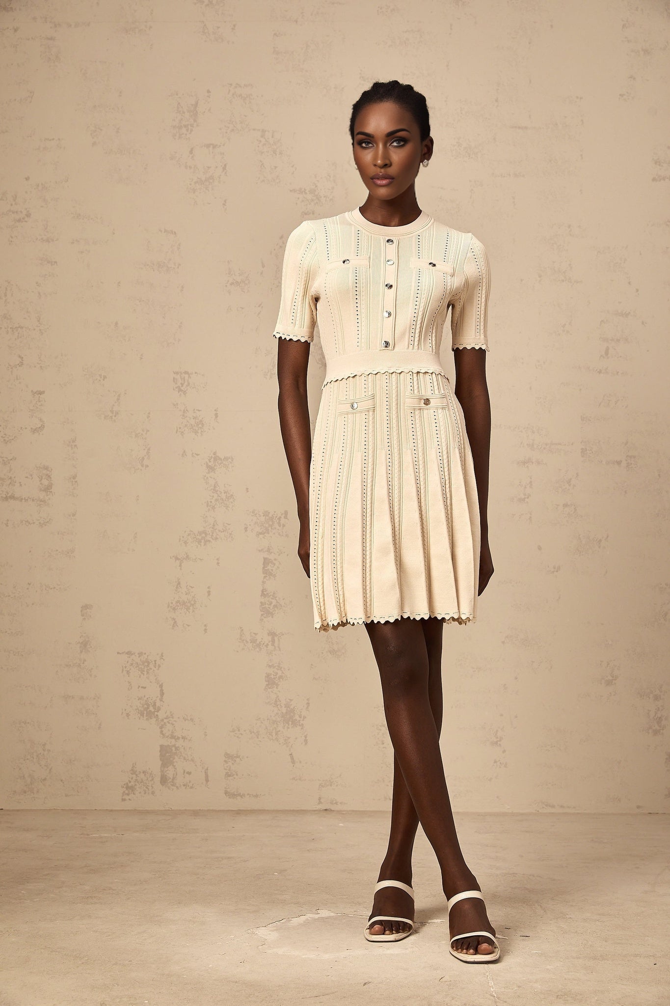 A woman wearing a short neutral colored knit dress with pleated skirt and delicate pointelle trim detailing