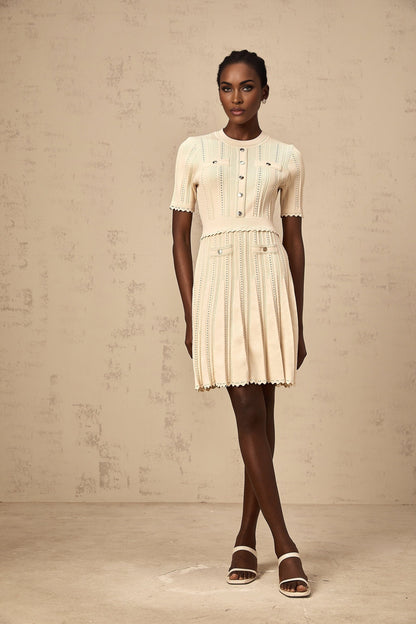 A woman wearing a short neutral colored knit dress with pleated skirt and delicate pointelle trim detailing