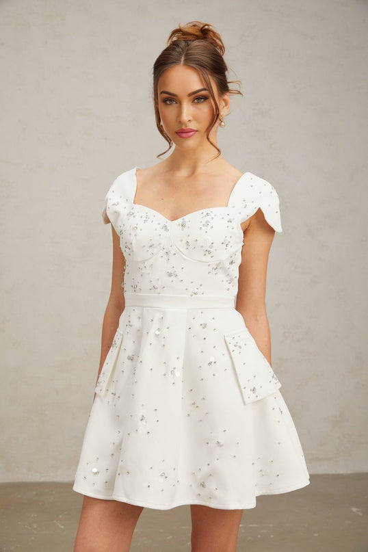 A woman wearing a short white flared dress with embellishments standing against a plain background