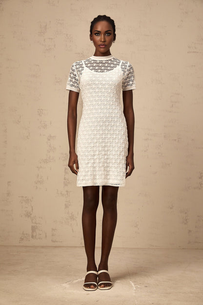 A woman wearing a short white semisheer knit dress standing in a neutral background
