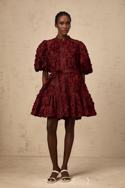 A woman wearing a short winered dress with three dimensional petal like puffs covering the fabric creating texture and volume