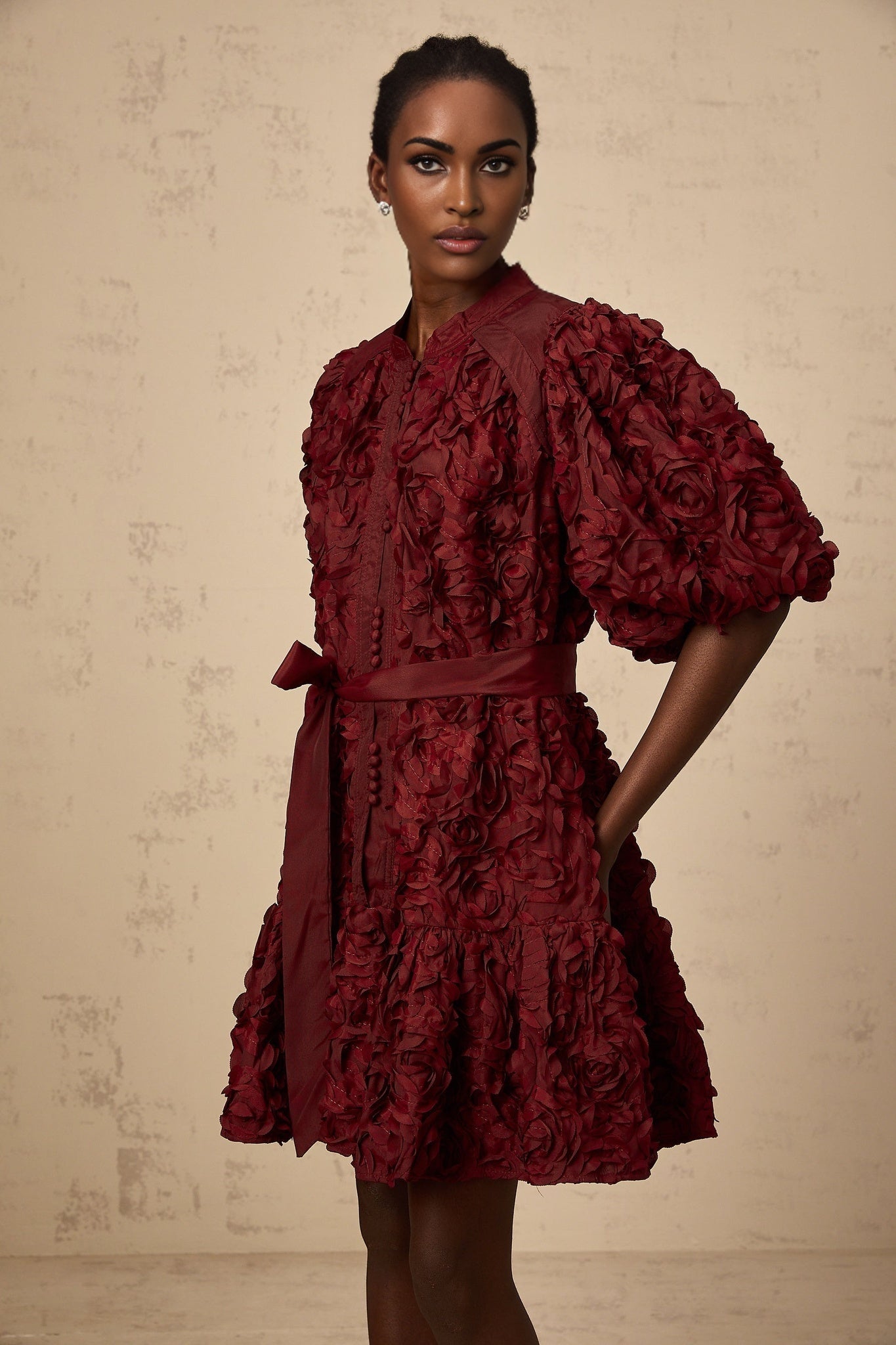 A woman wearing a short winered dress with three dimensional petallike puffs decorating the fabric standing against a plain background