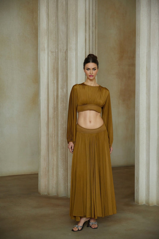 A woman wearing a vibrant green two piece outfit consisting of a fully pleated blouse and matching pleated skirt standing against a plain background