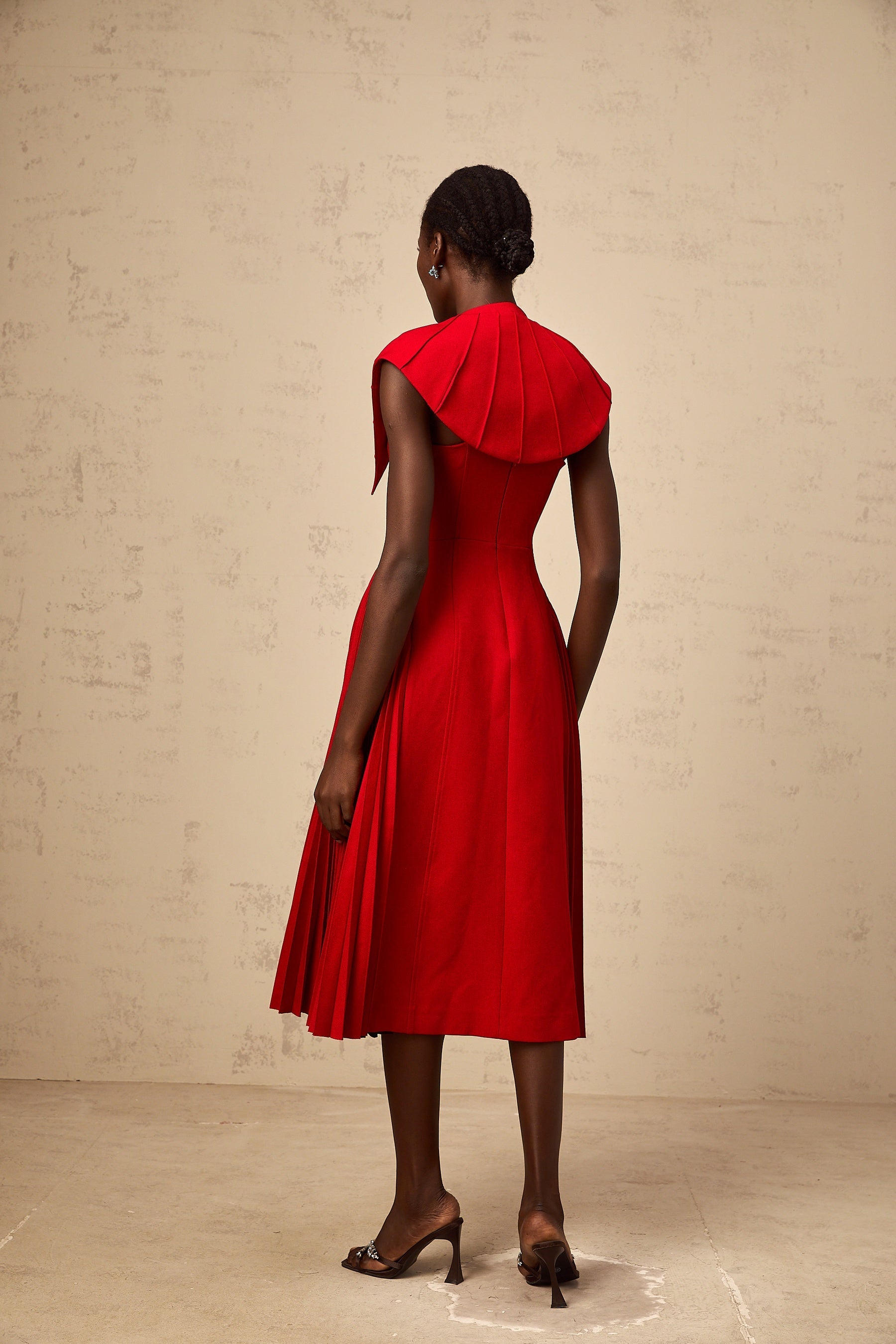 A woman wearing a vibrant red midi dress with a capelike design and pleated skirt standing in a neutral setting