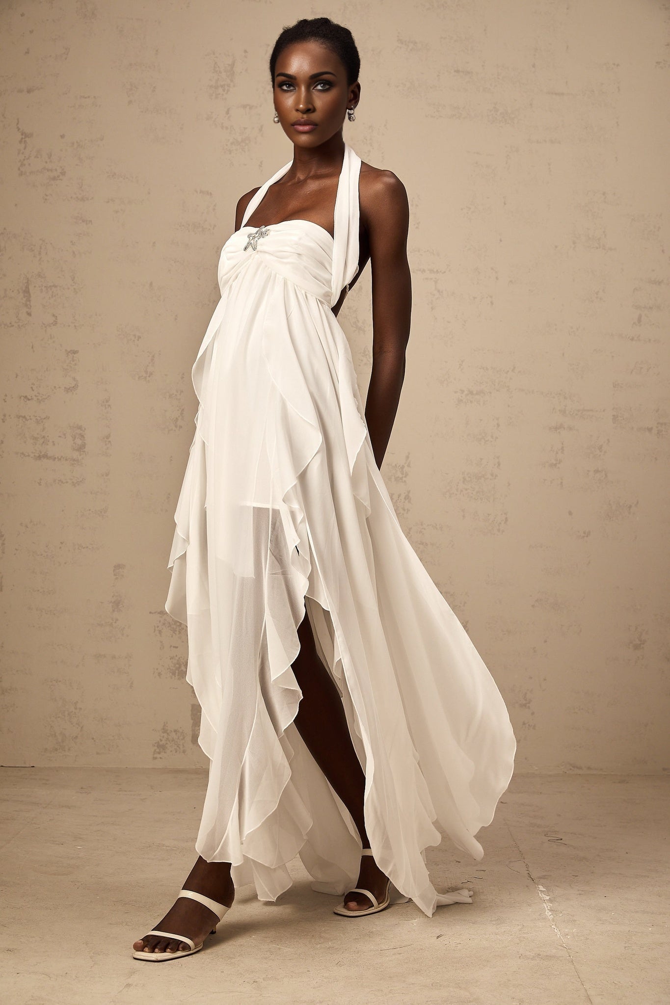 A woman wearing a white chiffon maxi dress with ruffles and a halter neck design standing against a plain background