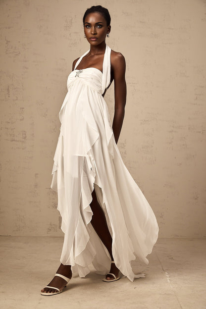 A woman wearing a white chiffon maxi dress with ruffles and a halter neck design standing against a plain background