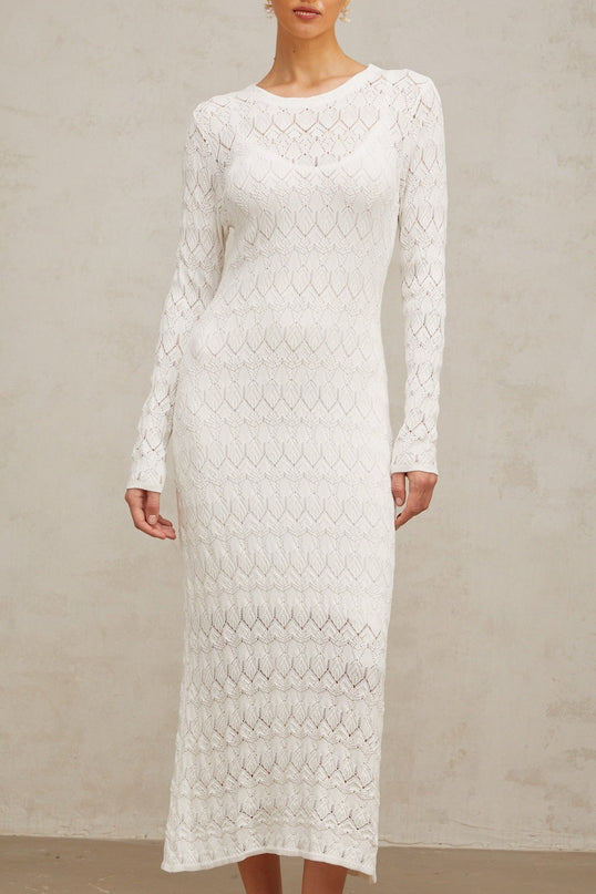 A woman wearing a white crochet knit midi dress with intricate lace patterns standing against a plain background