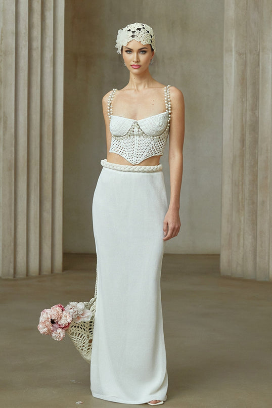 A woman wearing a white crochet knit two piece outfit consisting of a cropped top and midi skirt with delicate floral patterns standing against a plain background