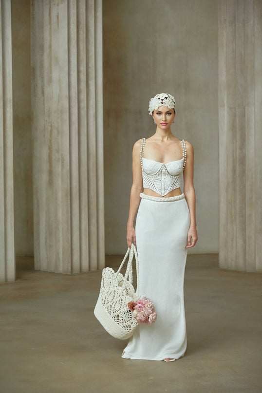 A woman wearing a white crochet knit two piece outfit consisting of a cropped top and midi skirt with floral pattern details standing against a plain background