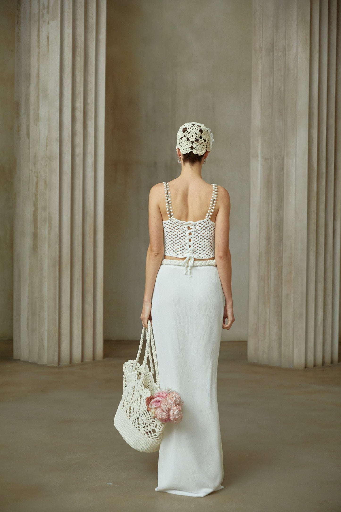 A woman wearing a white crochet knit two piece outfit consisting of a cropped top and midi skirt with intricate floral patterns standing against a plain background