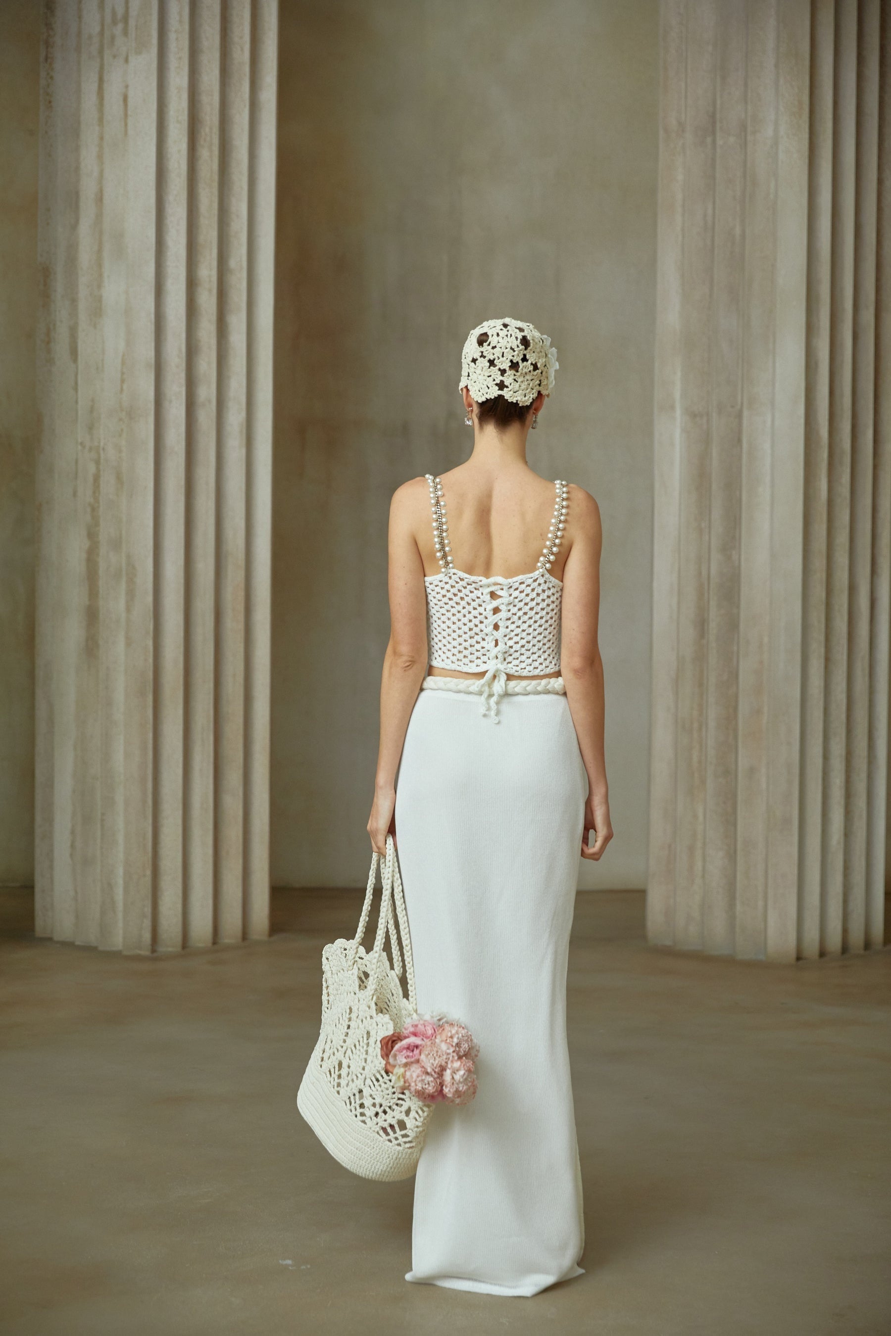 A woman wearing a white crochet knit two piece outfit consisting of a cropped top and midi skirt with intricate floral patterns standing against a plain background