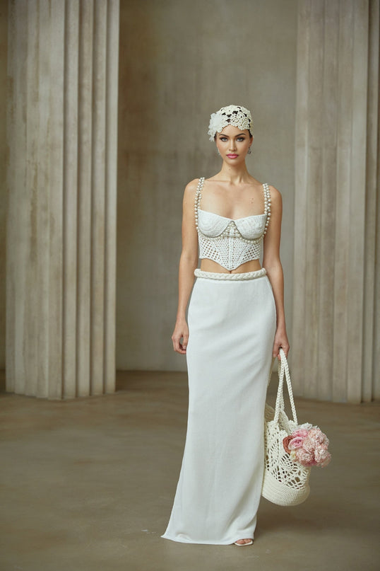 A woman wearing a white crochet knit two piece outfit consisting of a sleeveless crop top and a high waisted midi skirt with a floral pattern