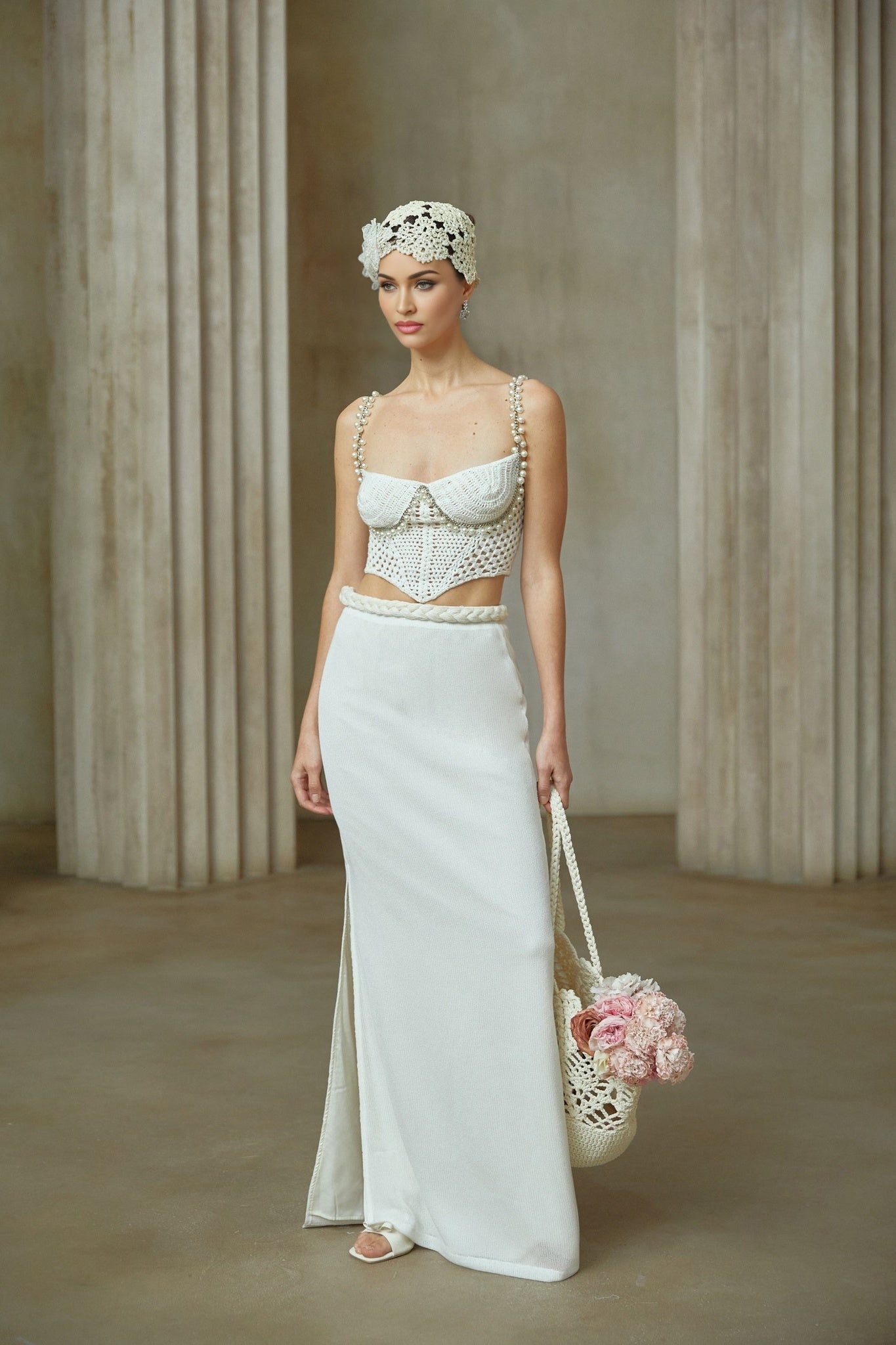 A woman wearing a white crochet knit two piece outfit consisting of a sleeveless crop top and matching high waisted midi skirt with a delicate floral pattern